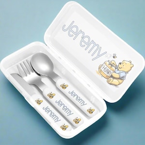 May include: A white plastic carrying case containing a metal fork, spoon, and knife set. Each utensil handle is white with the name Jeremy and a small bear graphic. The case lid features the name Jeremy and a bear holding a honey pot labelled HUNNY.