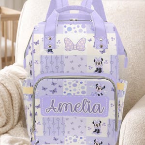 May include: A lavender and white diaper bag with a patchwork design featuring Minnie Mouse, butterflies, and hearts. The name "Amelia" is printed on the front. The bag has a top handle, shoulder straps, and multiple pockets.