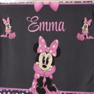 May include: A dark gray fleece blanket featuring a large Minnie Mouse graphic in the center, two smaller Minnie Mouse figures on the sides, and the name Emma printed in pink script. The blanket has a pink bow at the top and white trim.