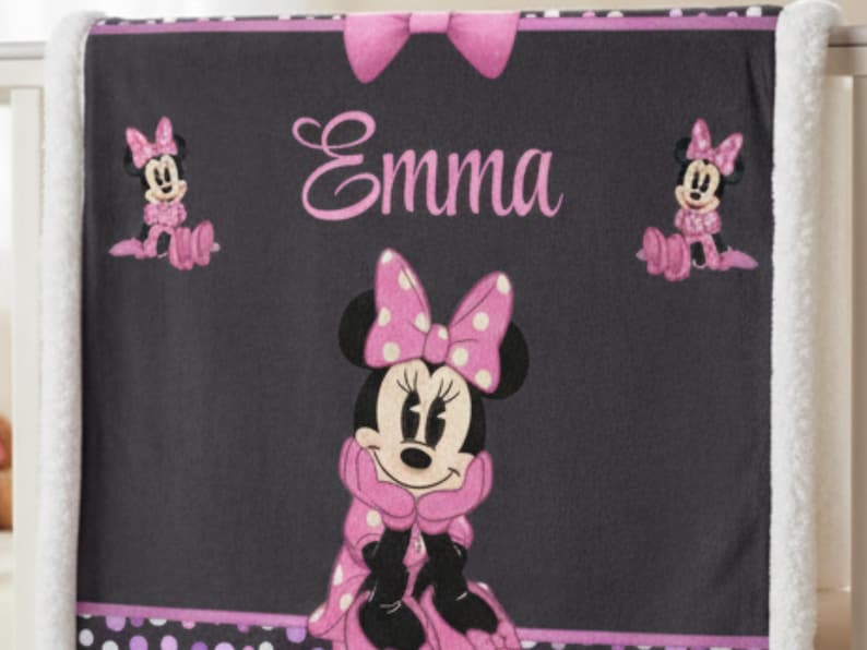 May include: A dark gray fleece blanket featuring a large Minnie Mouse graphic in the center, two smaller Minnie Mouse figures on the sides, and the name Emma printed in pink script. The blanket has a pink bow at the top and white trim.