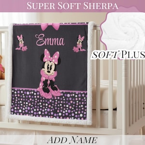 May include: A black and white sherpa baby blanket featuring a central graphic of Minnie Mouse in a pink polka dot dress and bow. The blanket includes the name Emma in pink script, a pink bow at the top, and a purple and pink polka dot border.