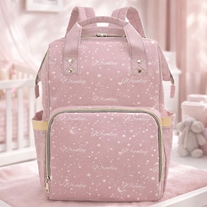 Puede incluir: Mochila rosa para bebé con el nombre "Wrenley" impreso en blanco, junto con estrellas y lunas. La bolsa tiene múltiples bolsillos, un asa superior y correas ajustables. La bolsa está sobre un cambiador rosa.
