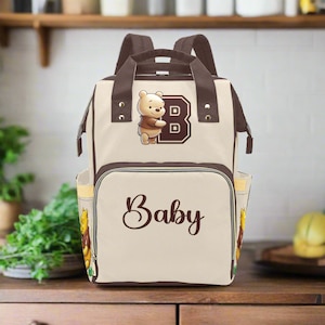 May include: A beige diaper bag with brown straps and a Winnie the Pooh character design. The bag has a large pocket on the front with the word "Baby" printed in brown cursive lettering. The bag also has a smaller pocket on the front with a Winnie the Pooh character and the letter "B" printed in brown.