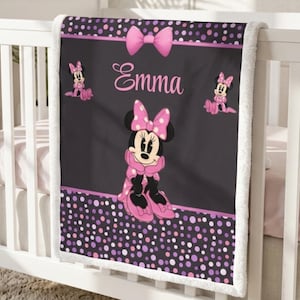 May include: A black and pink fleece blanket featuring a central illustration of Minnie Mouse, the name Emma in pink script, a pink bow at the top, and smaller Minnie Mouse figures on the sides. The border has a pattern of pink and purple polka dots.