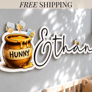 May include: Wooden wall decor featuring the name "Ethan" in a cursive font, accompanied by a honey pot with the word "HUNNY" and cartoon bees. The honey pot is brown and gold, with honey dripping down. The bees have blue wings.