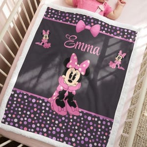 May include: A rectangular baby blanket with a white sherpa border, featuring a dark gray center with a Minnie Mouse graphic, a pink bow, and the name Emma in pink script. The top and bottom borders have a purple, pink, and white polka dot pattern.
