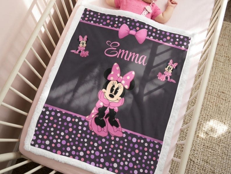 May include: A rectangular baby blanket with a white sherpa border, featuring a dark gray center with a Minnie Mouse graphic, a pink bow, and the name Emma in pink script. The top and bottom borders have a purple, pink, and white polka dot pattern.