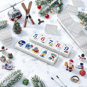 May include: A set of white mahjong tiles featuring winter and holiday-themed illustrations, including numbers 5 through 9, a ski helmet, gloves, a sled, a lantern, and a scarf, arranged on a snowy surface with miniature holiday decorations.