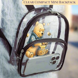 May include: Clear compact mini backpack featuring Winnie the Pooh with a honey pot and bees. The clear backpack has a black trim, a zippered main compartment, and a front pocket holding a smartphone. The text "CLEAR COMPACT MINI BACKPACK" is at the top.