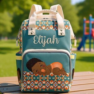May include: Teal diaper bag with the name "Elijah" in white script. The front pocket features a cartoon baby illustration. The bag has cream-colored handles and trim, with a geometric pattern in orange, brown, and cream.
