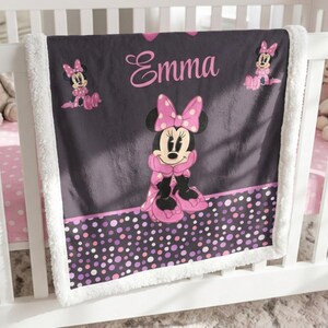 May include: A dark gray fleece blanket with a white sherpa border featuring a central graphic of Minnie Mouse in a pink polka dot bow and dress. The name Emma is printed in pink script above the character, with smaller Minnie Mouse icons and a bottom border of multicolored pink and purple polka dots.