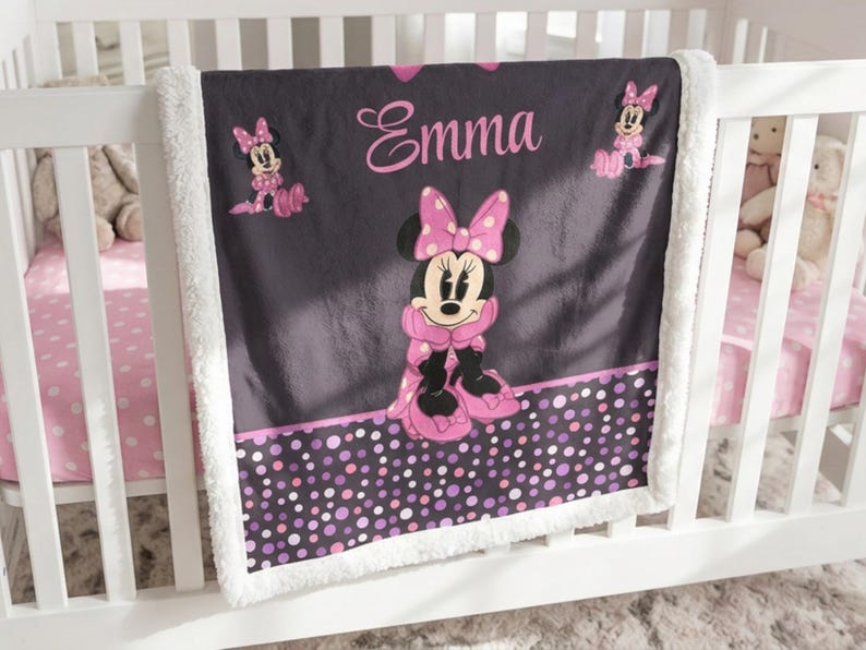 May include: A dark gray fleece blanket with a white sherpa border featuring a central graphic of Minnie Mouse in a pink polka dot bow and dress. The name Emma is printed in pink script above the character, with smaller Minnie Mouse icons and a bottom border of multicolored pink and purple polka dots.