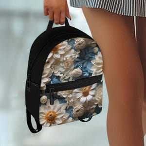 May include: A small black backpack featuring a front panel with a detailed floral pattern. The design displays white daisy-like flowers with yellow centers and smaller spherical white blossoms set against a muted blue background with foliage.