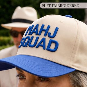 May include: A beige baseball cap with a blue brim and the text "MAHJ SQUAD" in blue puff embroidery on the front. The cap features a structured crown with a blue button on top and matching blue stitching along the brim and panels.