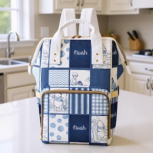 May include: A blue and white patchwork backpack with a Winnie the Pooh theme. The backpack features the name "Noah" embroidered on two navy blue squares. It has white straps and a top handle, with gold-coloured zips and accents. The design includes polka dots, stripes, and gingham patterns.