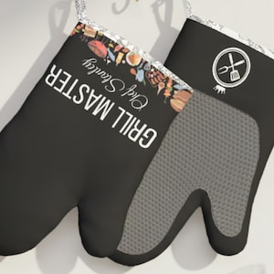 May include: Black oven mitts with the words "GRILL MASTER Chef Stanley" in white. One mitt features a grey, textured grip and a decorative border with food illustrations. The other mitt has a grill logo.