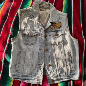 May include: Pale blue denim vest with a collared neckline, two chest pockets, and a button-up front. The vest has a "Candy Custom" patch and a small pin. The interior lining has a floral pattern.
