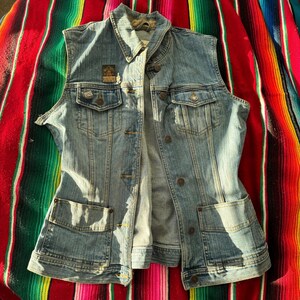 Puede incluir: Un chaleco vaquero azul claro con cuello, cierre de botones y múltiples bolsillos. El chaleco tiene un aspecto descolorido y presenta pequeños parches en el pecho. El chaleco es sin mangas y parece de estilo vintage.