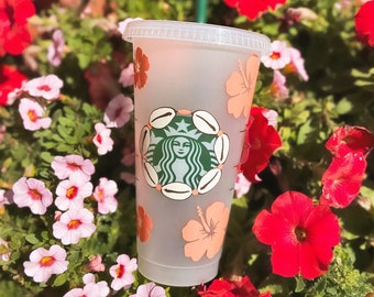 May include: A clear plastic reusable cup with a pink and green floral design. The cup has a white lid and a straw. The design features a mermaid, seashells, and flowers.