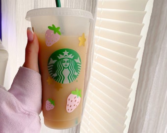May include: A clear plastic cup with a green straw and a Starbucks logo. The cup has a pink and green strawberry design and gold stars.