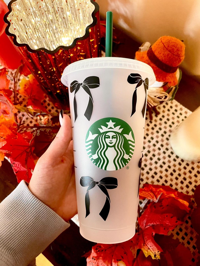 May include: A clear, reusable cold cup with a black bow pattern and a green Starbucks logo. The cup has a green straw and a white lid. The cup is held in front of a blurred background with fall-themed decorations.