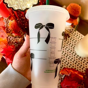 May include: A clear plastic cold beverage cup with a black bow design. The cup has a green straw and markings for drink customization. The cup is held by a person.