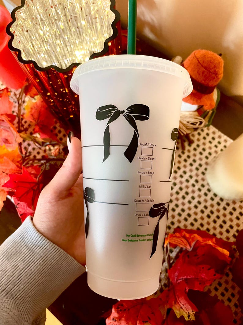 May include: A clear plastic cold beverage cup with a black bow design. The cup has a green straw and markings for drink customization. The cup is held by a person.