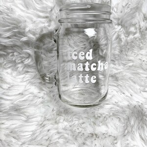 May include: A clear glass mason jar with a handle, featuring the text "iced matcha latte" in white lettering.