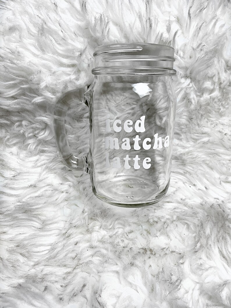 May include: A clear glass mason jar with a handle, featuring the text "iced matcha latte" in white lettering.
