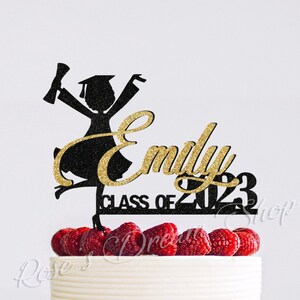 May include: Black silhouette of a graduate with a gold glitter name "Emily" and the text "Class of 2023" on a white cake with red raspberries.