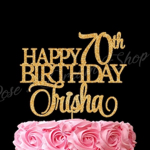 May include: A pink frosted cake with a gold glitter cake topper that reads "Happy Birthday Trisha 70th". The cake is decorated with pink frosting roses and sits on a white cake stand against a black background.