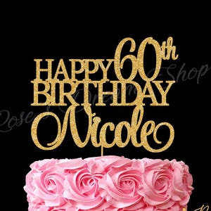 May include: A pink frosted cake with a gold glitter cake topper that reads "Happy 60th Birthday Nicole." The cake is on a white cake stand against a black background. The cake has a rose design.