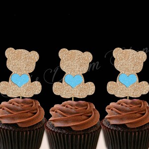 May include: Three chocolate cupcakes with brown frosting topped with glitter teddy bear cupcake toppers. Each teddy bear is holding a light blue heart.