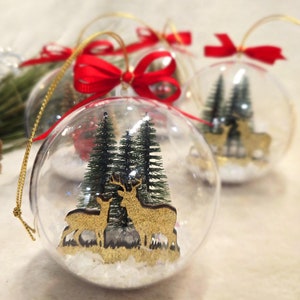 May include: Clear glass Christmas baubles with a snowy scene inside. The baubles feature two gold reindeer and small green trees. Each bauble has a red ribbon bow and a gold string for hanging.