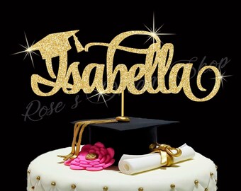 May include: Gold glitter graduation cake topper with the name "Isabella" and a graduation cap. The topper is on a white cake with pink and gold decorations.