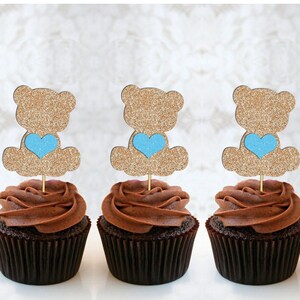 May include: Three chocolate cupcakes with gold glitter teddy bear toppers holding blue hearts. The cupcakes are frosted with chocolate frosting.