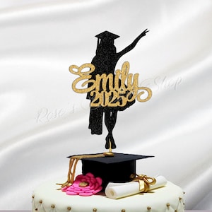 May include: A white cake decorated with pink roses and gold accents. A black graduation cap and a diploma are on top of the cake. A black silhouette of a graduate with the name "Emily 2023" in gold glitter is on top of the cap.