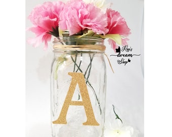 May include: A clear glass mason jar featuring a large, glittery gold letter A on the front. The jar is wrapped with natural raffia twine around the neck and contains a bouquet of pink carnations. The text Rosie's dream Shop is visible.
