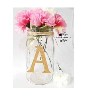 May include: A clear glass mason jar with a gold glitter letter A on the front. The jar is filled with pink and white carnations and tied with a brown twine bow. The text "Rosie's dream shop" is written in black on a white background.
