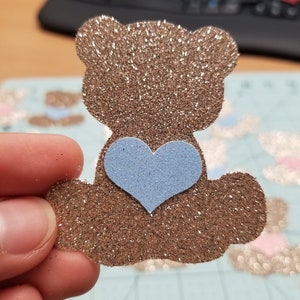 May include: A brown glitter teddy bear with a light blue glitter heart on its chest.