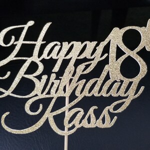 Pode incluir: Topo de bolo de glitter dourado com o texto "Happy 18th Birthday Kass" em fonte cursiva.