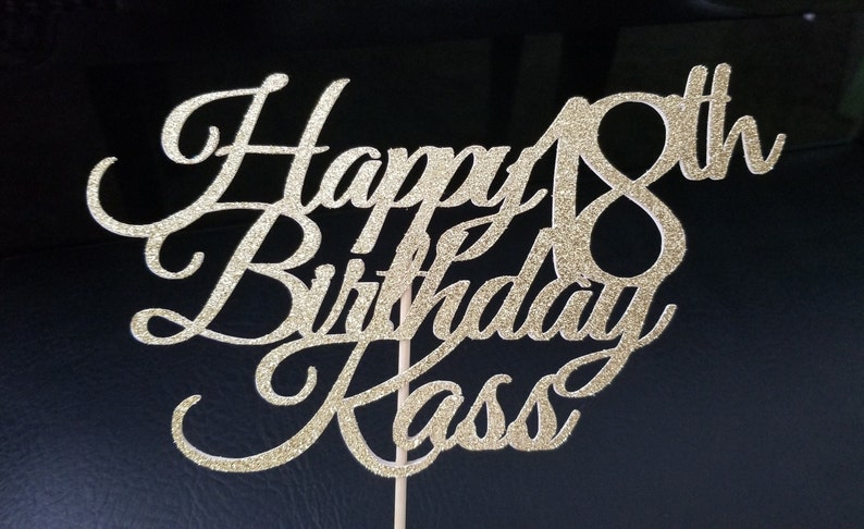 Pode incluir: Topo de bolo de glitter dourado com o texto "Happy 18th Birthday Kass" em fonte cursiva.