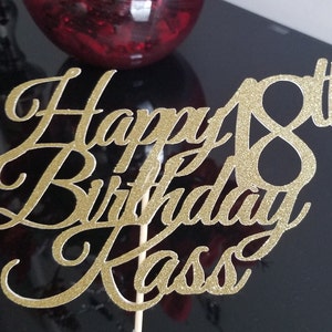 Pode incluir: Topo de bolo de glitter dourado com o texto "Happy 18th Birthday Kass" em fonte cursiva.