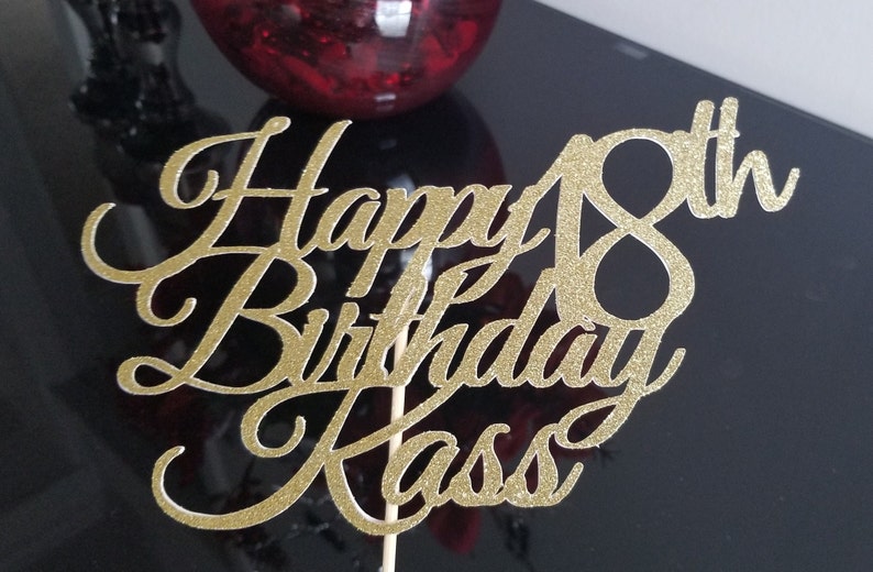Pode incluir: Topo de bolo de glitter dourado com o texto "Happy 18th Birthday Kass" em fonte cursiva.