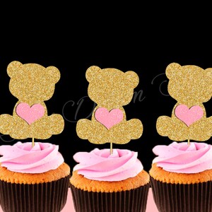 May include: Three cupcakes with pink frosting and gold glitter teddy bear toppers holding pink hearts. The teddy bears are sitting down.