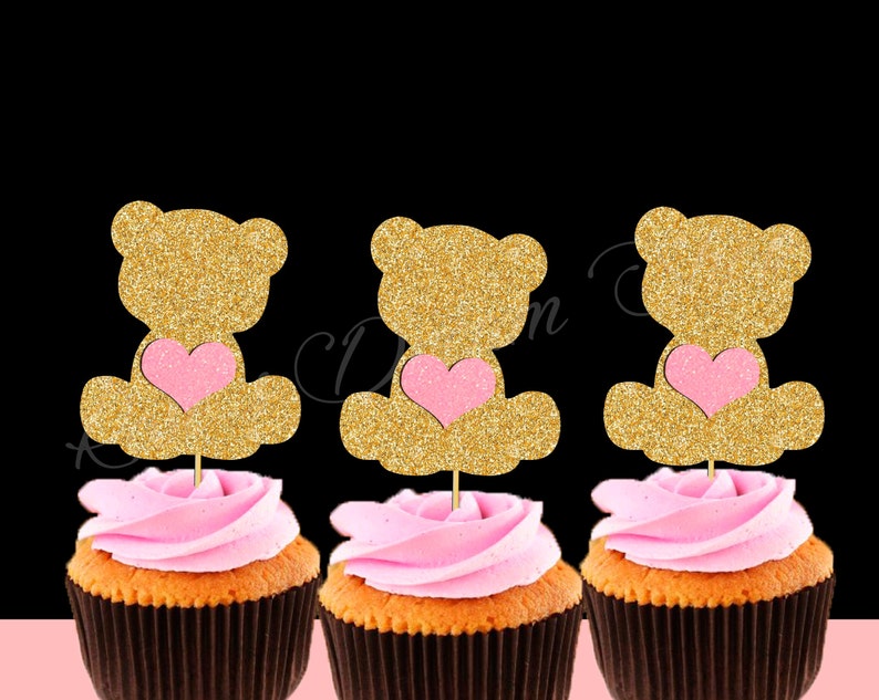 May include: Three cupcakes with pink frosting and gold glitter teddy bear toppers holding pink hearts. The teddy bears are sitting down.