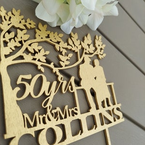 May include: Gold-colored wooden cake topper with a tree silhouette, two figures, and the text "50 yrs Mr & Mrs COLLINS".