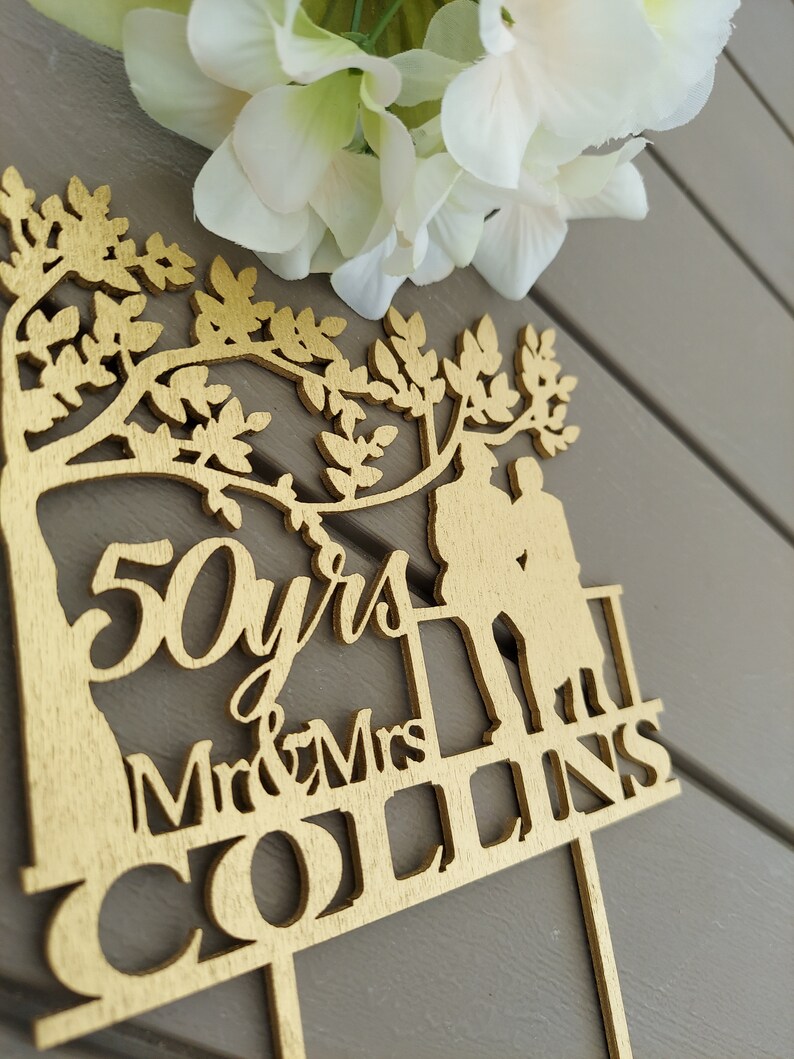 May include: Gold-colored wooden cake topper with a tree silhouette, two figures, and the text "50 yrs Mr & Mrs COLLINS".
