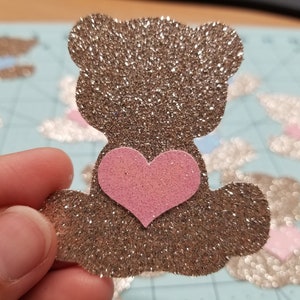 May include: A brown glitter teddy bear with a pink glitter heart on its chest. The bear is holding a pink glitter heart in its paws.