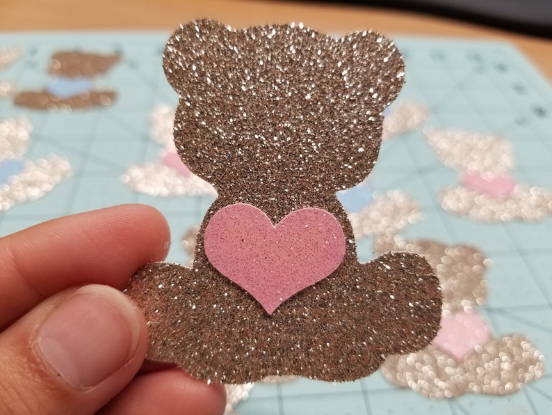 May include: A brown glitter teddy bear with a pink glitter heart on its chest. The bear is holding a pink glitter heart in its paws.